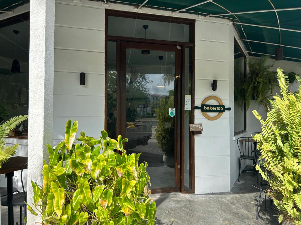 Bakerloo's entrance under a green awning, with a round timber logo by the glass door and ferns either side