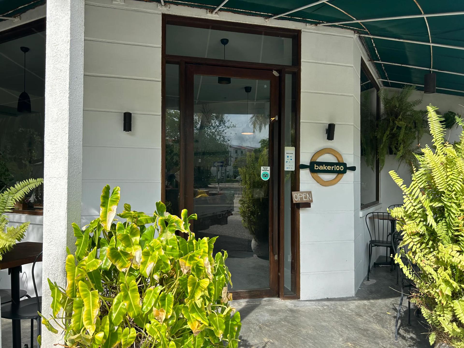 Bakerloo's entrance under a green awning, with a round timber logo by the glass door and ferns either side