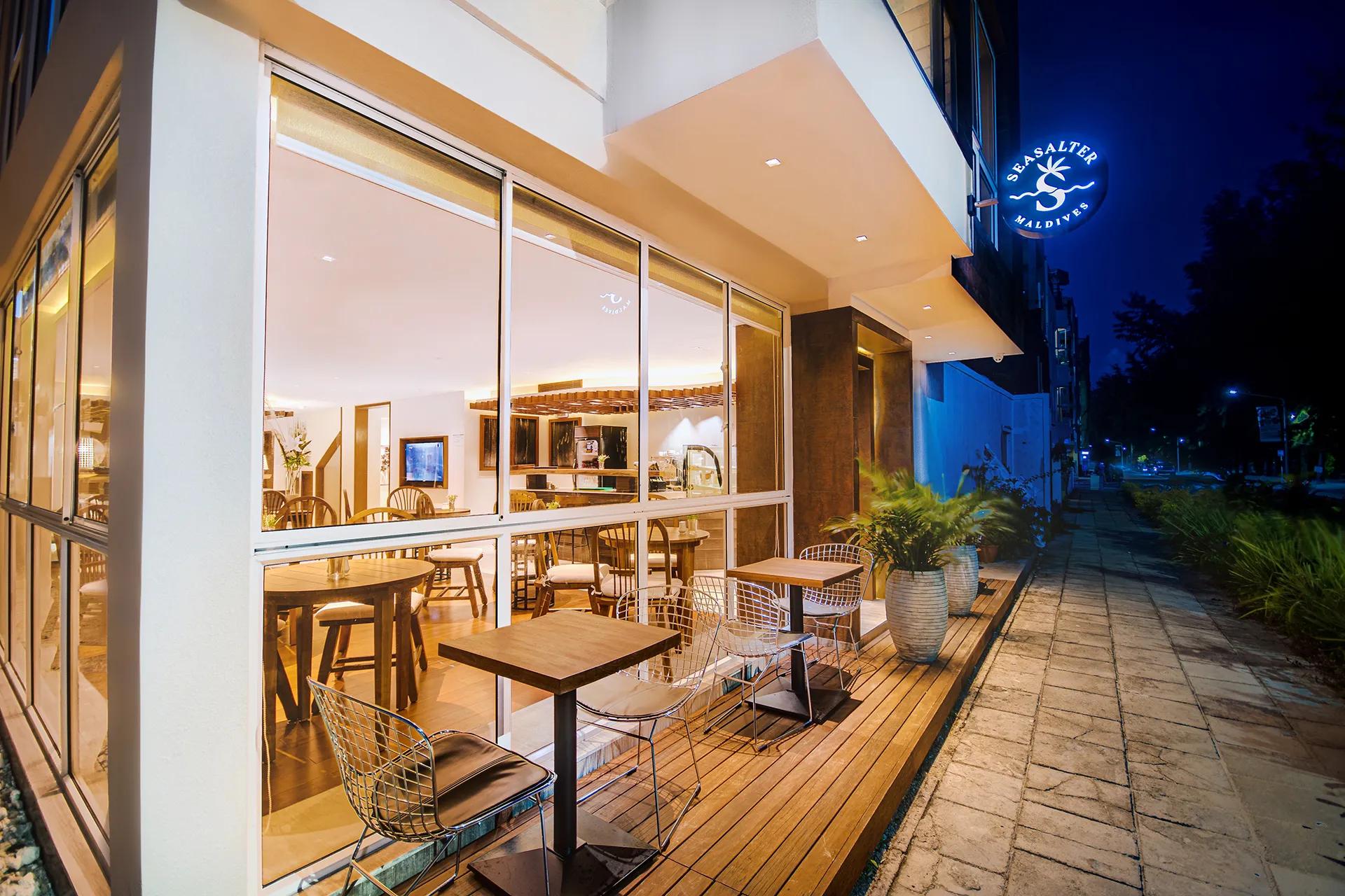 Evening exterior with café terrace seating and the lit Seasalter Maldives sign at the entrance in Hulhumalé.