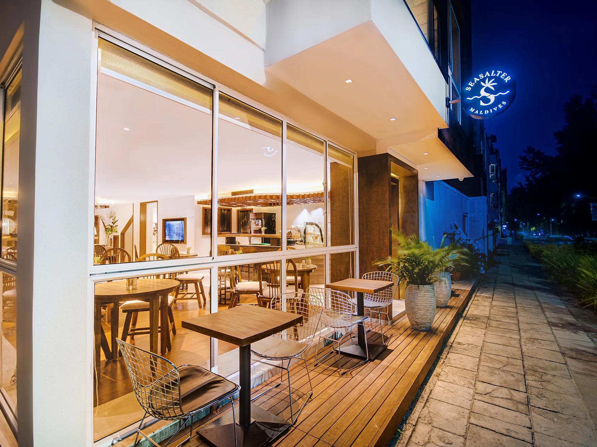 Evening exterior with café terrace seating and the lit Seasalter Maldives sign at the entrance in Hulhumalé.