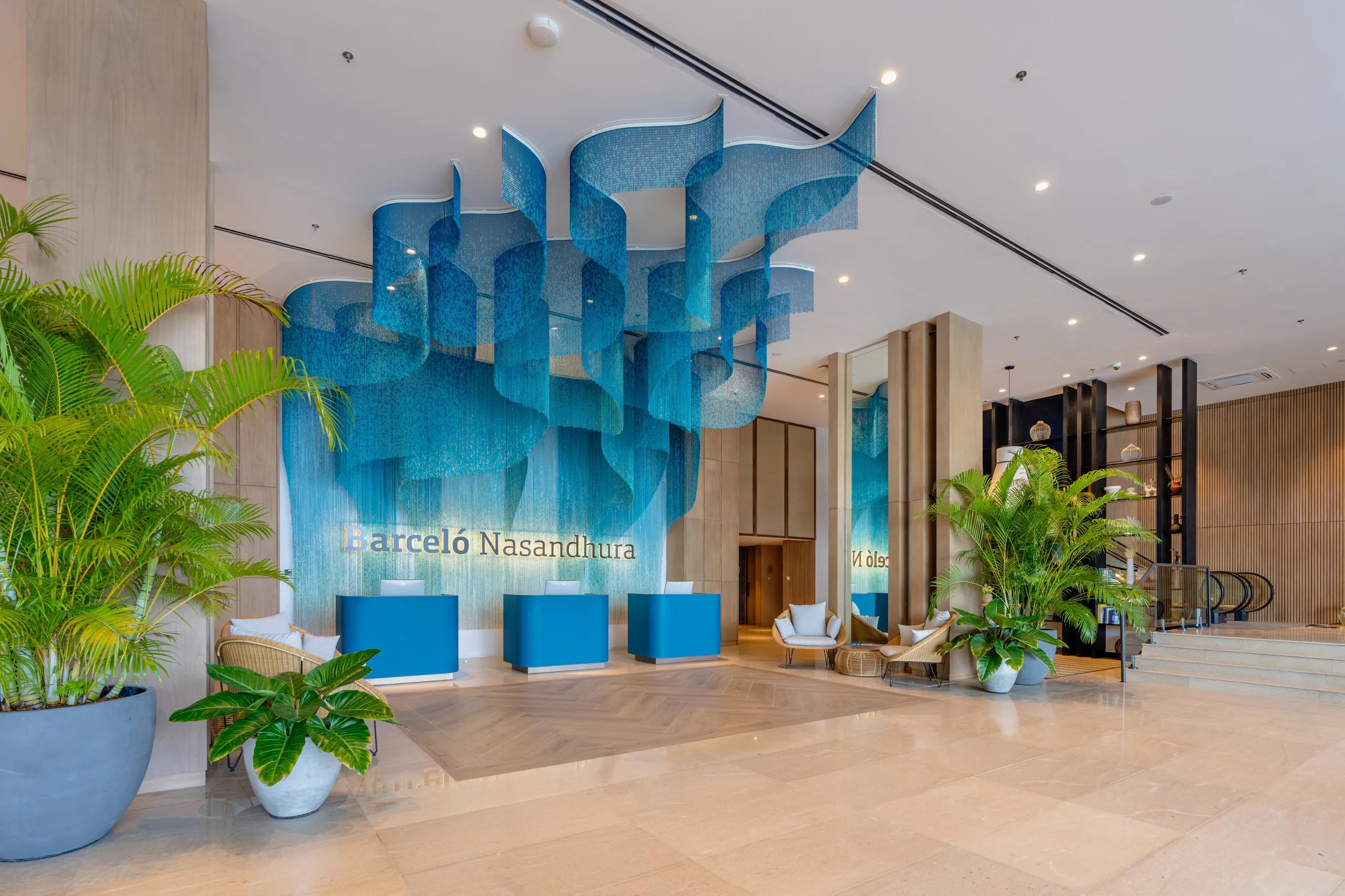 Hotel lobby with three bright blue reception desks below a cascading blue bead installation and the illuminated Barceló Nasandhura name on the wall