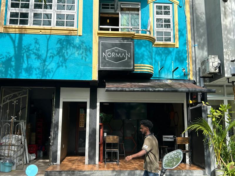 The Norman sign on a turquoise building, with bar stools at the open shopfront and a man walking past