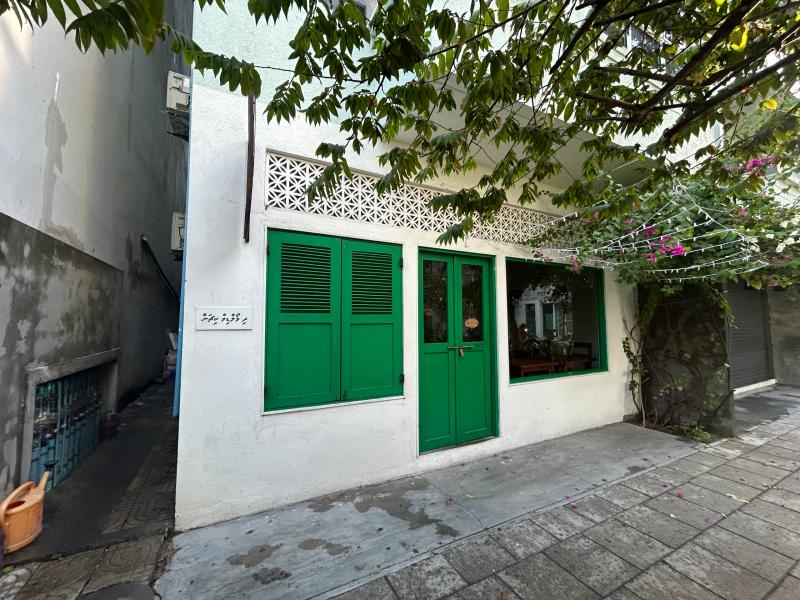 Whitewashed building with green louvred doors and a Dhivehi sign, set on a paved lane with bougainvillea overhead