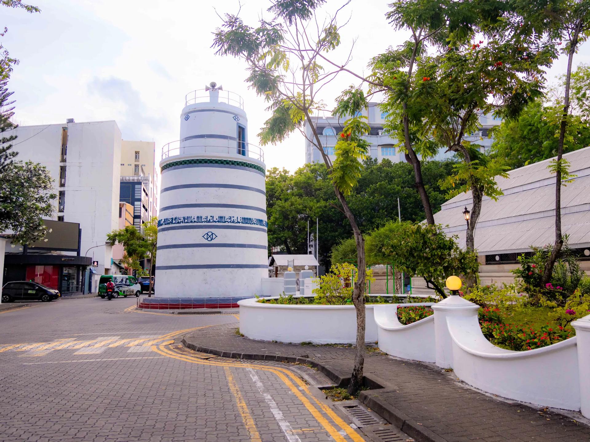 White cylindrical minaret with blue Arabic inscription beside Hukuru Miskiy, on a paved street corner