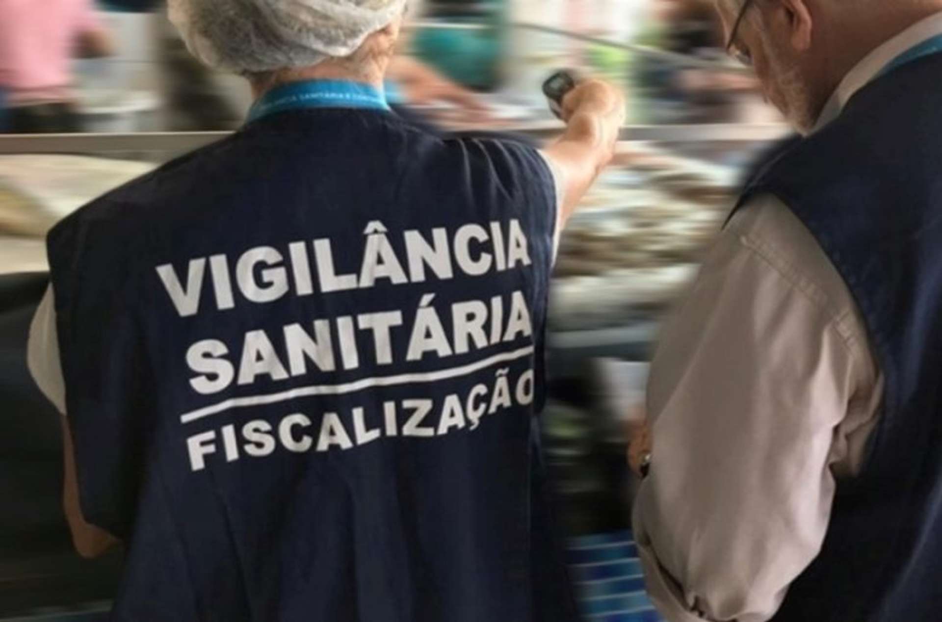 Agentes da Vigilância Sanitária realizam fiscalização em estabelecimento, usando coletes azuis com a inscrição “Vigilância Sanitária – Fiscalização”, enquanto observam e inspecionam o local.