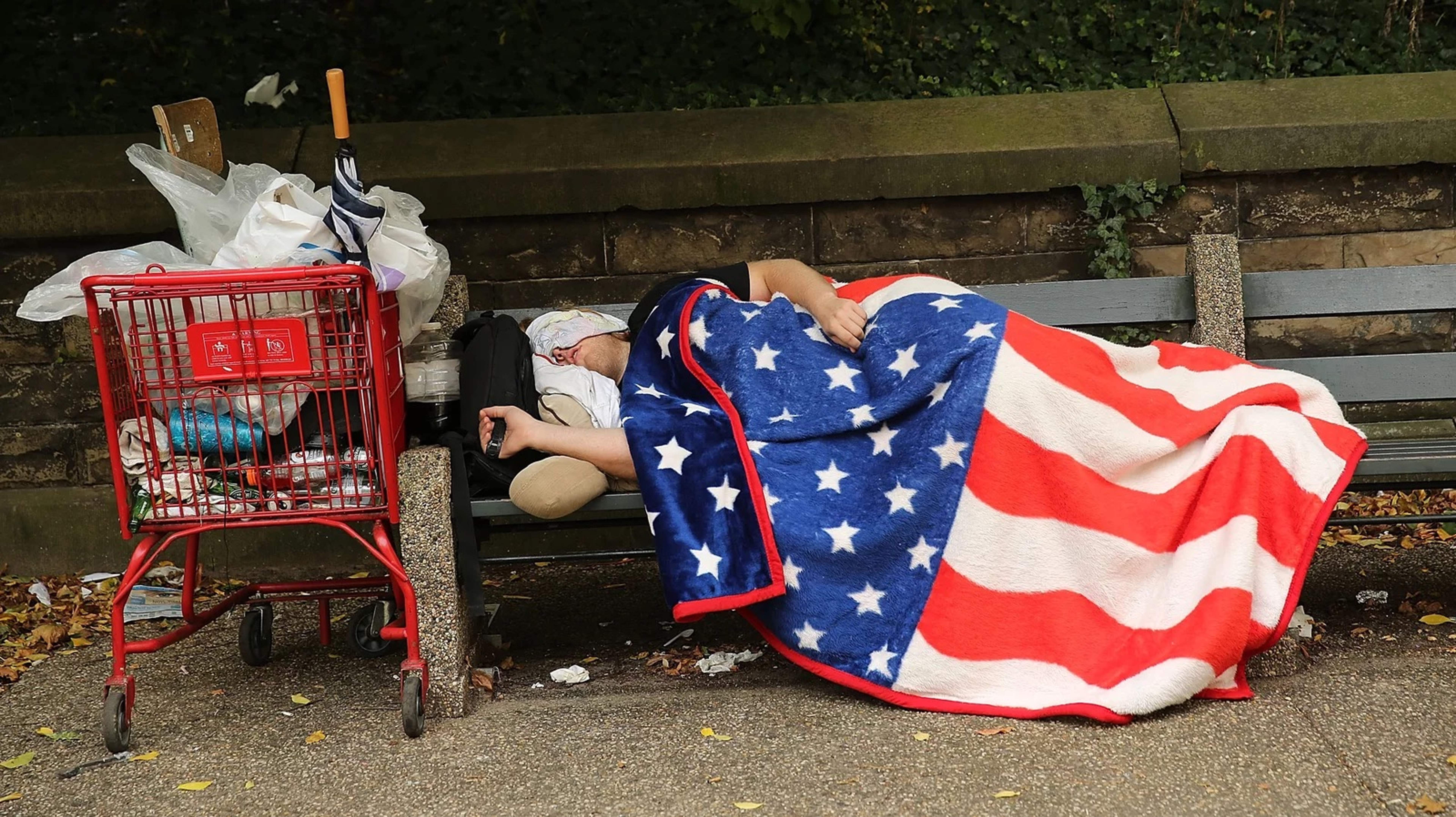 Unhoused person sleeping on a bench with an American flag blanket.