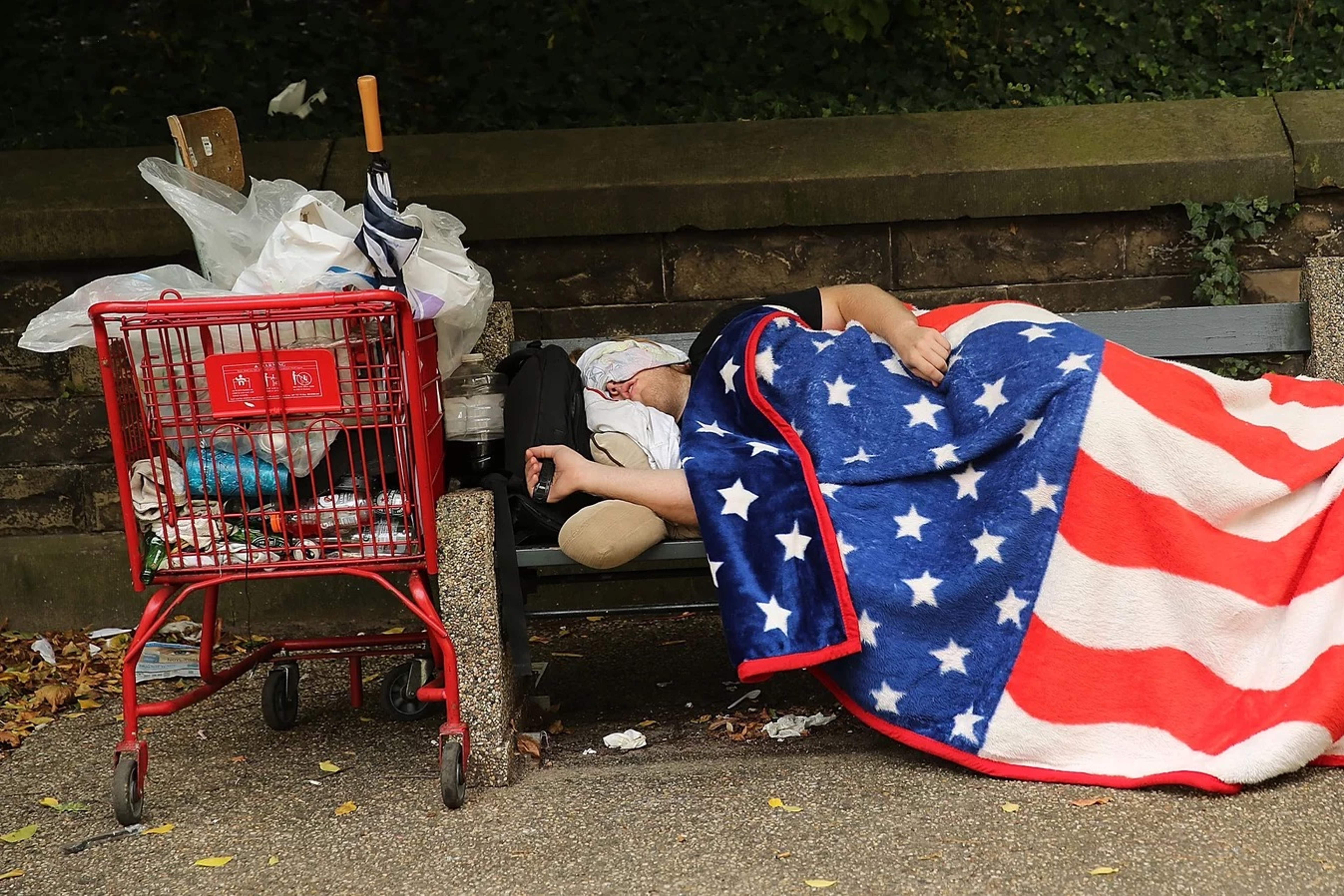Unhoused person sleeping on a bench with an American flag blanket.