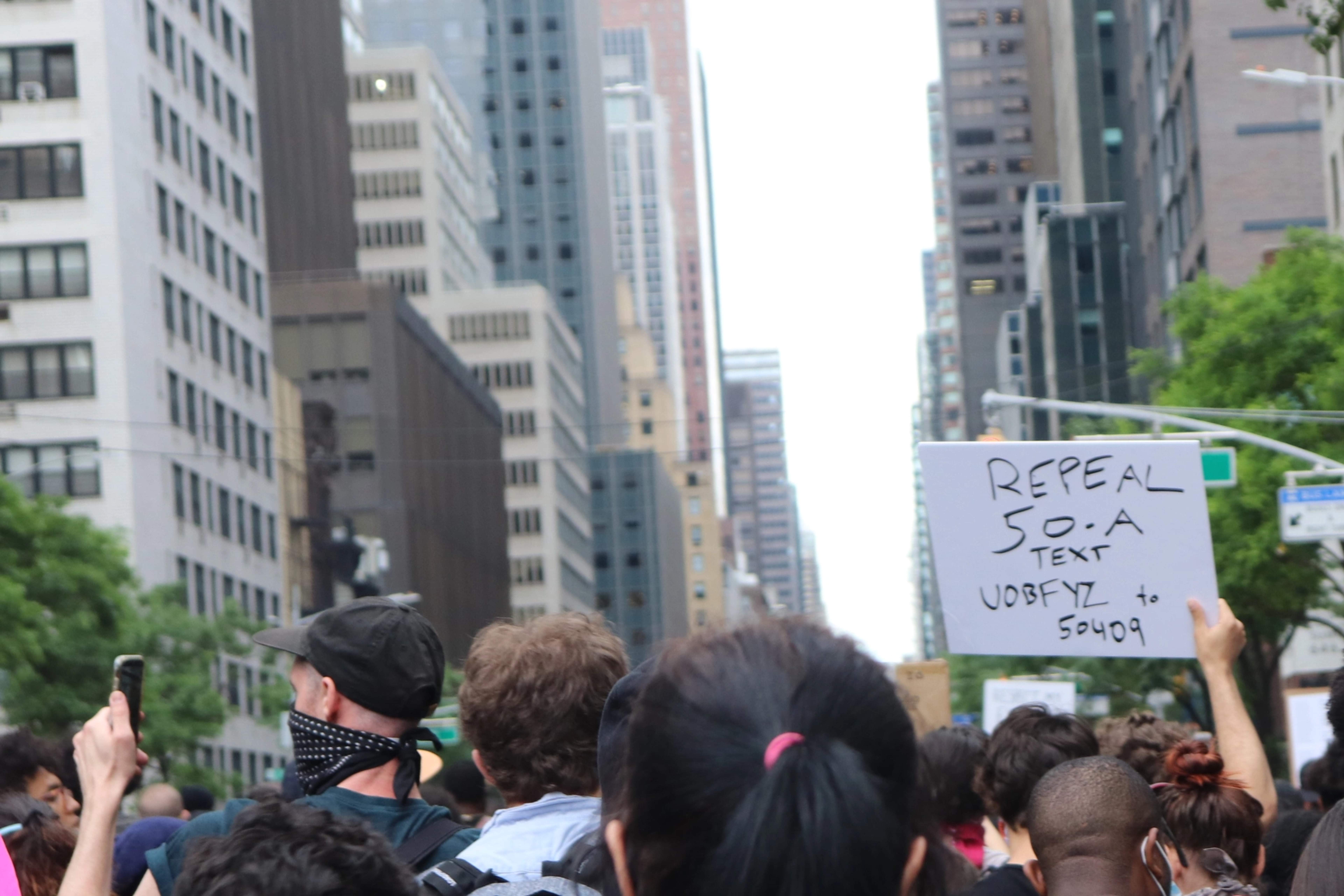 Crowded protest in New York City featuring a protestor holding a sign that reads "Text UOBFYZ to 50409"