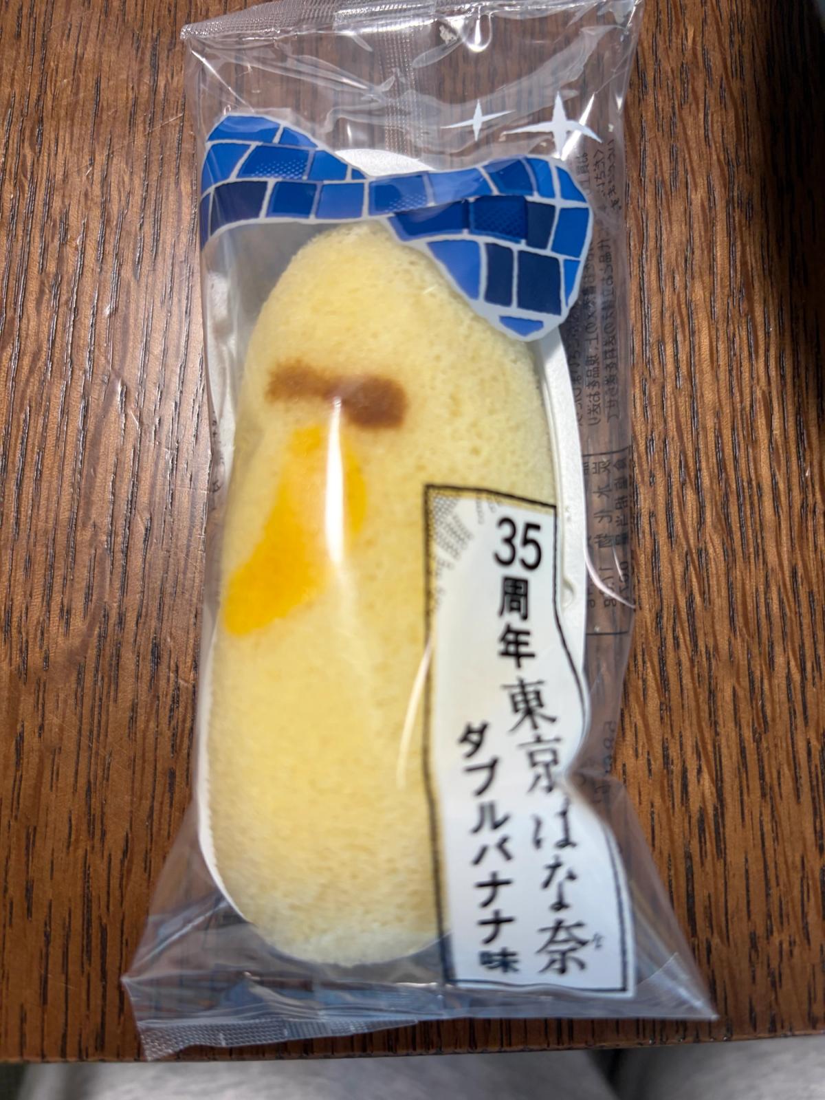 個包装の東京ばな奈。透明の袋に青いミラーボール柄のリボンと「35周年東京ばな奈 ダブルバナナ味」のラベルが印刷され、中のスポンジケーキにバナナの焼き印が見える。