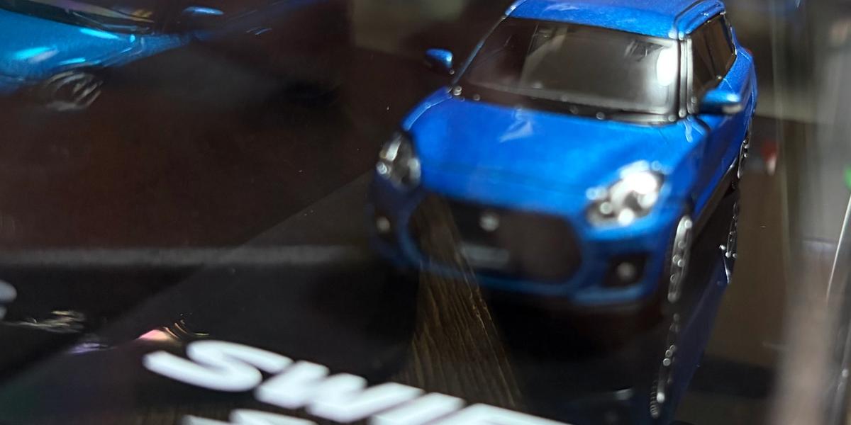 クリアケースに入った Hobby JAPAN 1/64 スイフトスポーツ ZC33S を斜め前から見たところ。青い車体と、SWIFT SPORT のロゴが入った黒い台座が見える