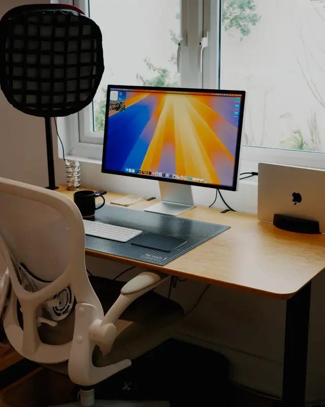A desk by a window with a large monitor, a laptop on a stand and a white chair