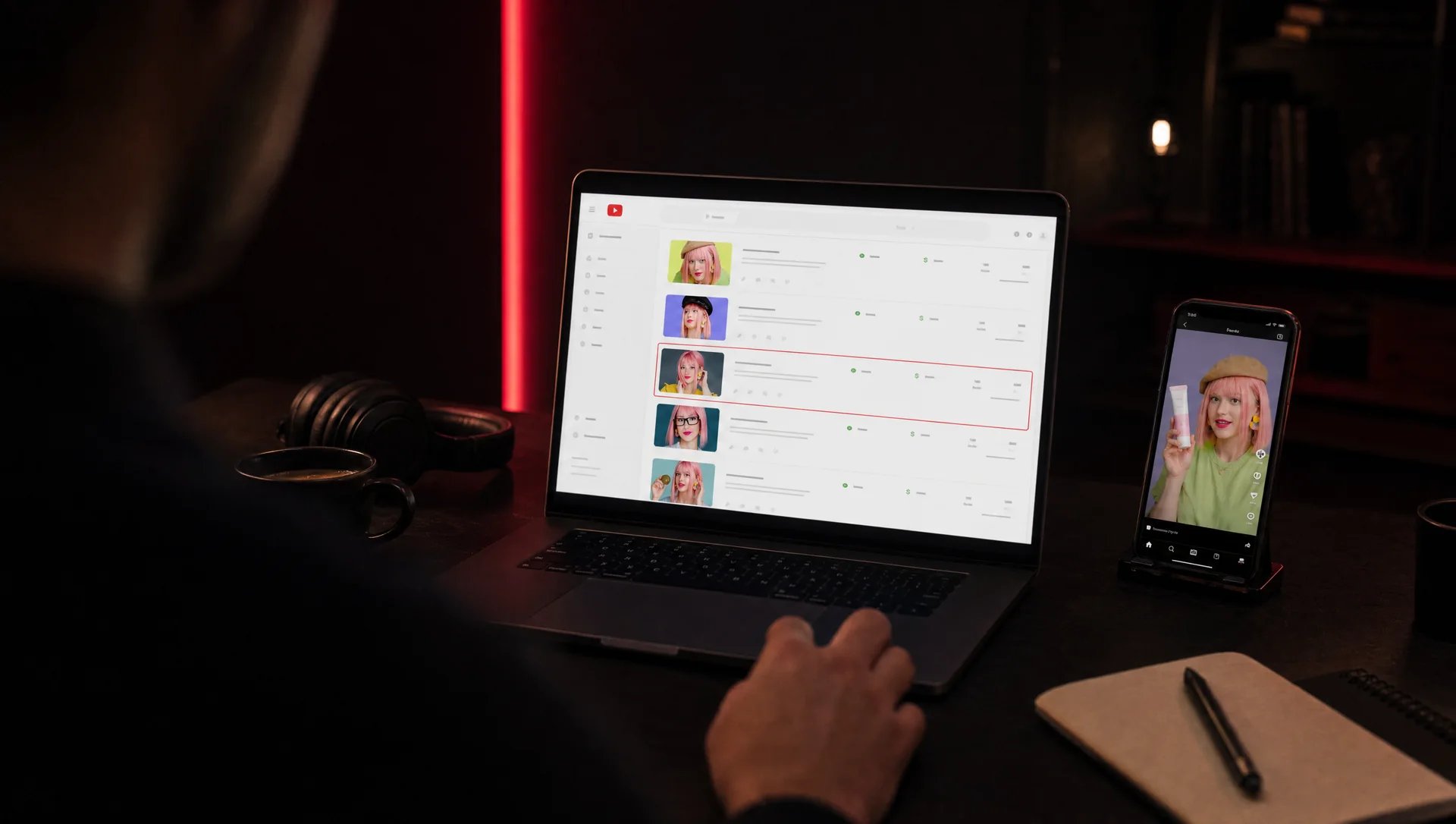حاسوب محمول يعرض في YouTube Studio قائمة فيديوهات يظهر فيها الوجه نفسه، وأحد الأسطر محاط بإطار أحمر، وهاتف يعرض إعلانًا مزيفًا بهذا الوجه، في استوديو مظلم بإضاءة حمراء