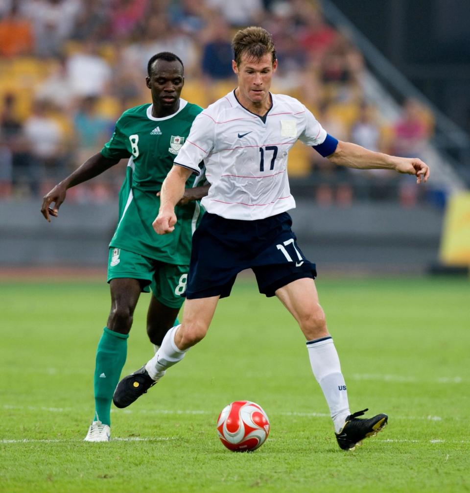 brian mcbride in white jersey and dark shorts dribbling a ball in front of an opponent