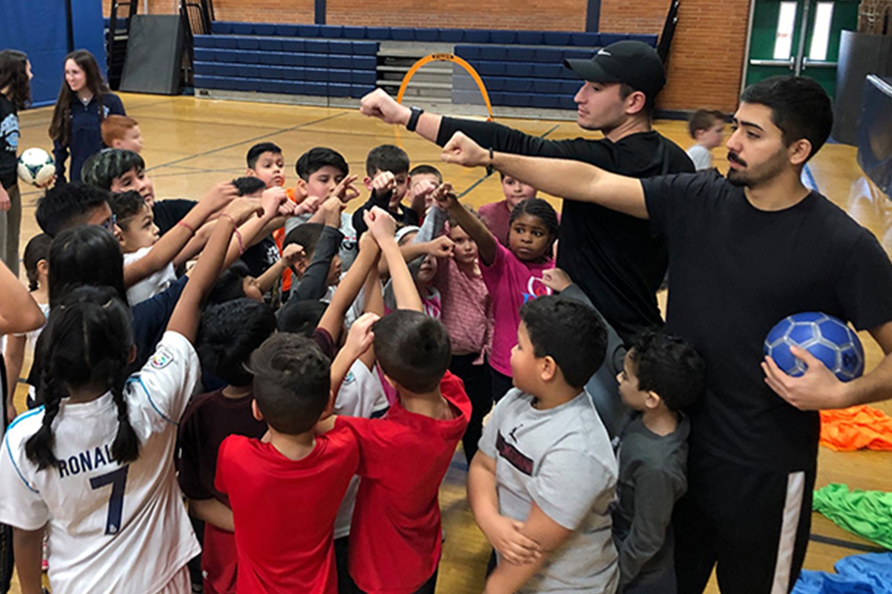 Youth soccer coaches lead a team huddle inside a school gym, with children raising their hands together before a soccer activity.