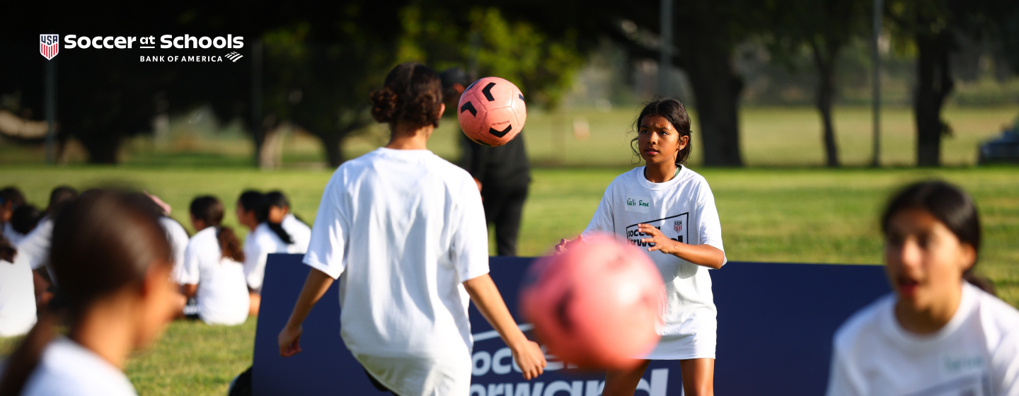 Two girls participate in a soccer activity at school, practicing ball control with a pink soccer ball on a grass field. Text on the image reads “Soccer at Schools.”