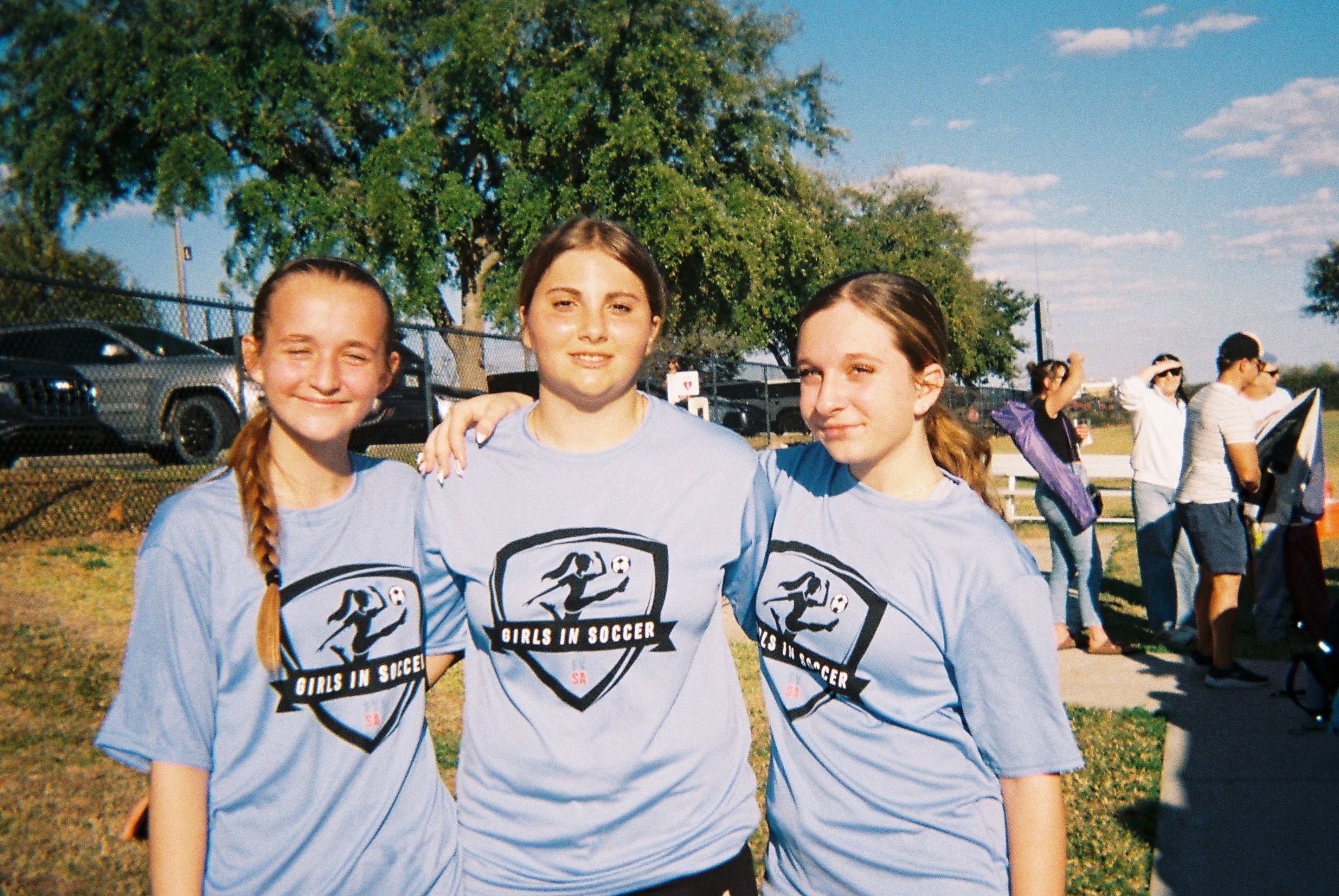 Three girls in matching “Girls in Soccer” shirts stand with arms around each other on a field, smiling at the camera.