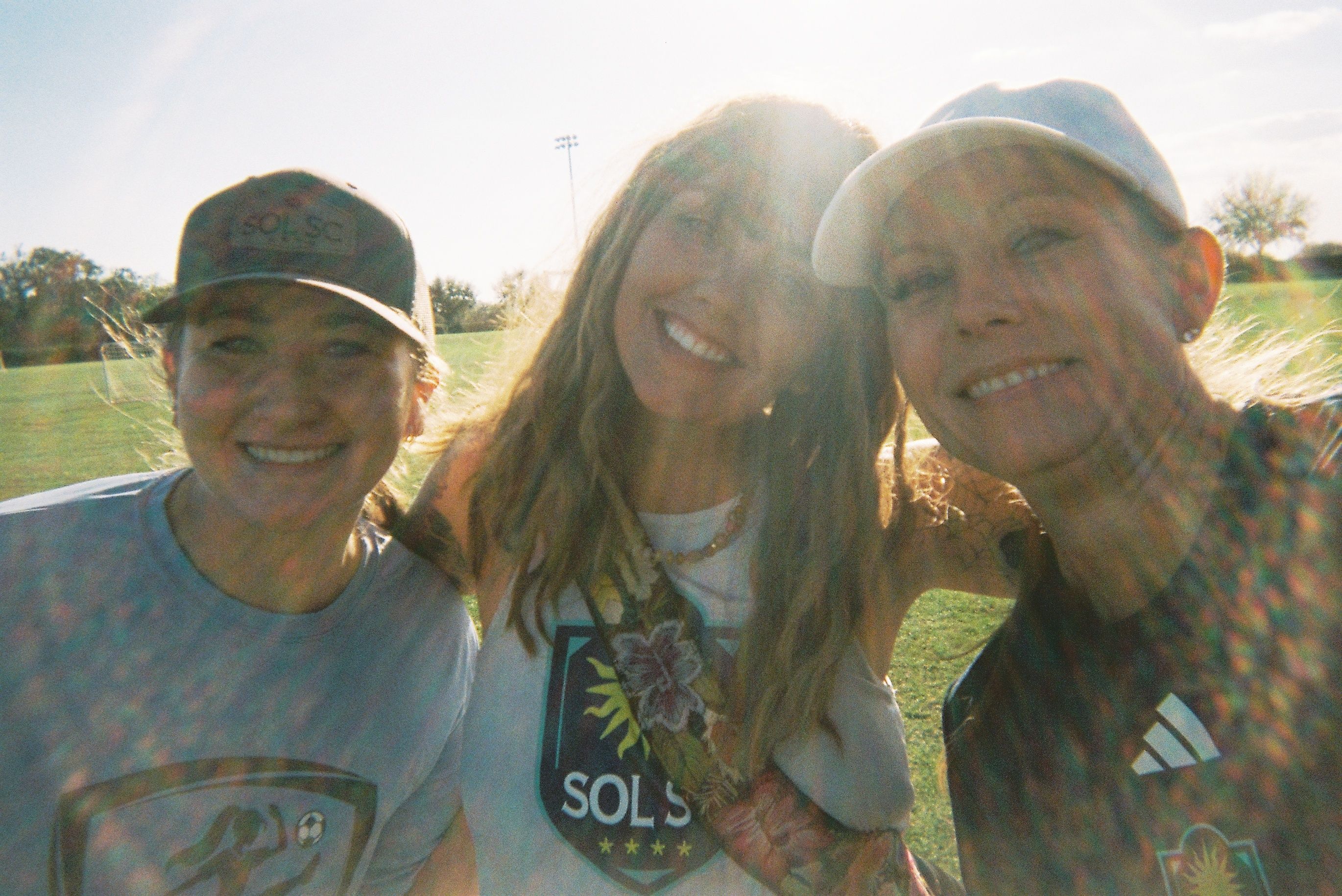Three women smiling together outdoors on a soccer field.