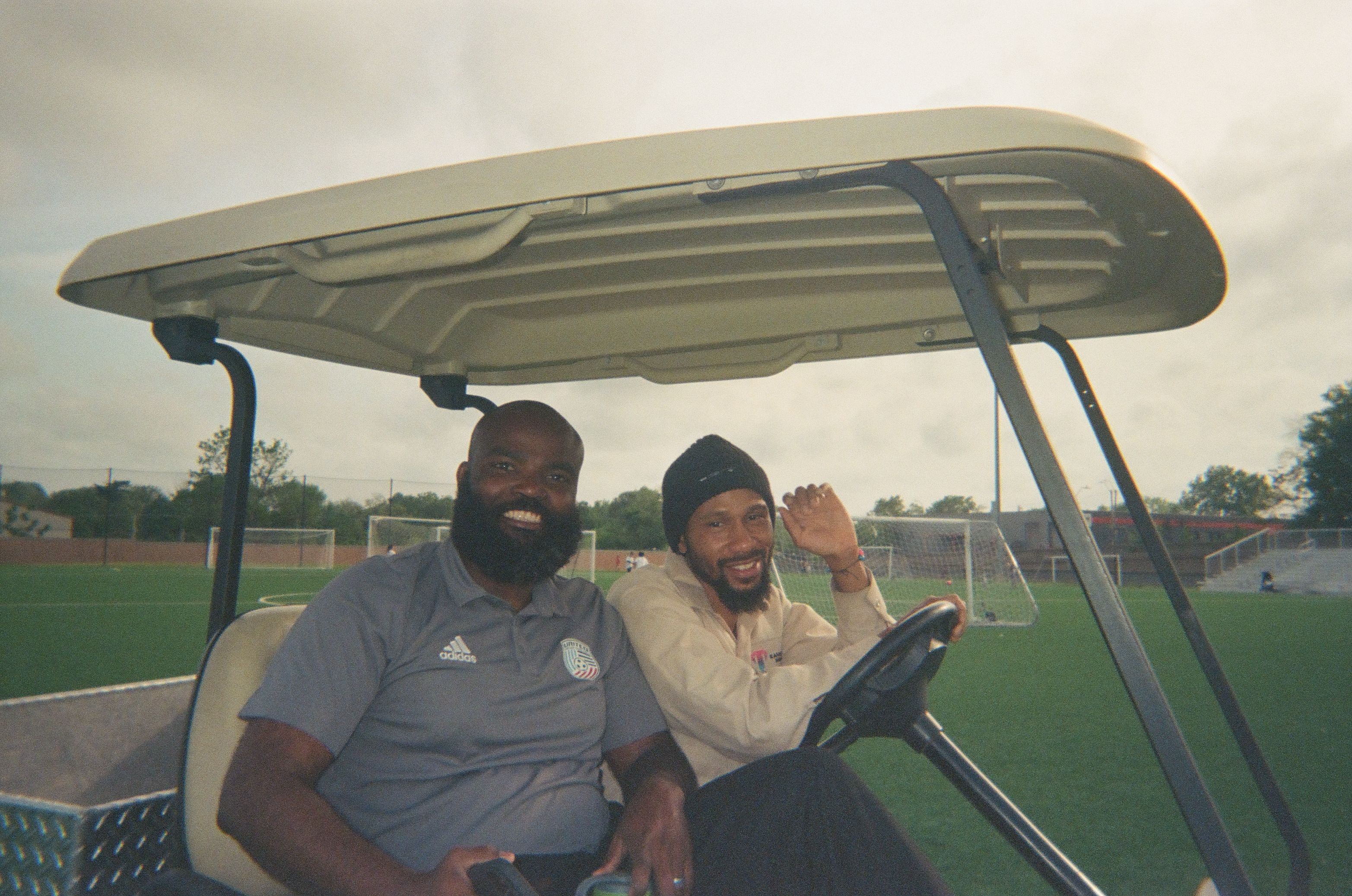 Two adults smile while sitting in a golf cart beside an outdoor soccer field.