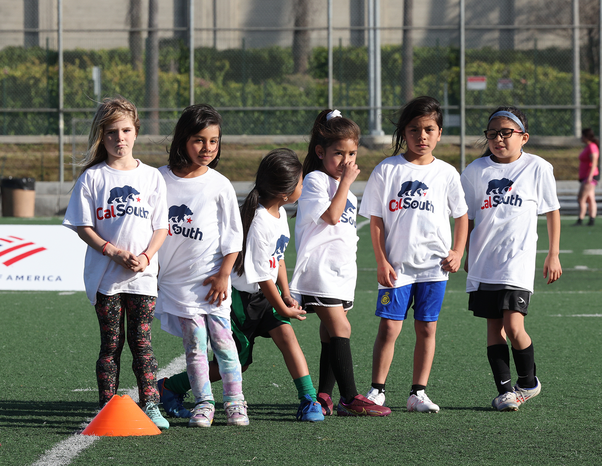 A group of young girls standing in a line on a soccer field during a youth practice, wearing white T-shirts, shorts, shin guards, and cleats, waiting near an orange cone.
