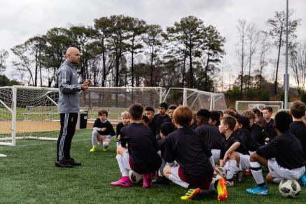 A group of young soccer players sits or kneels listening to instruction from a coach