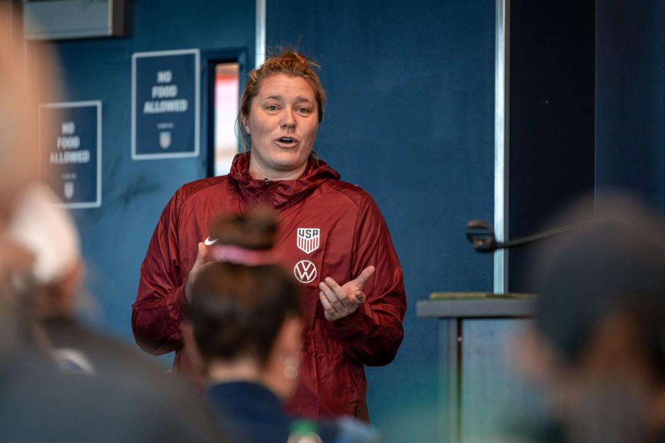 A women in a red jacket with a US Soccer crest addresses a seated crowd