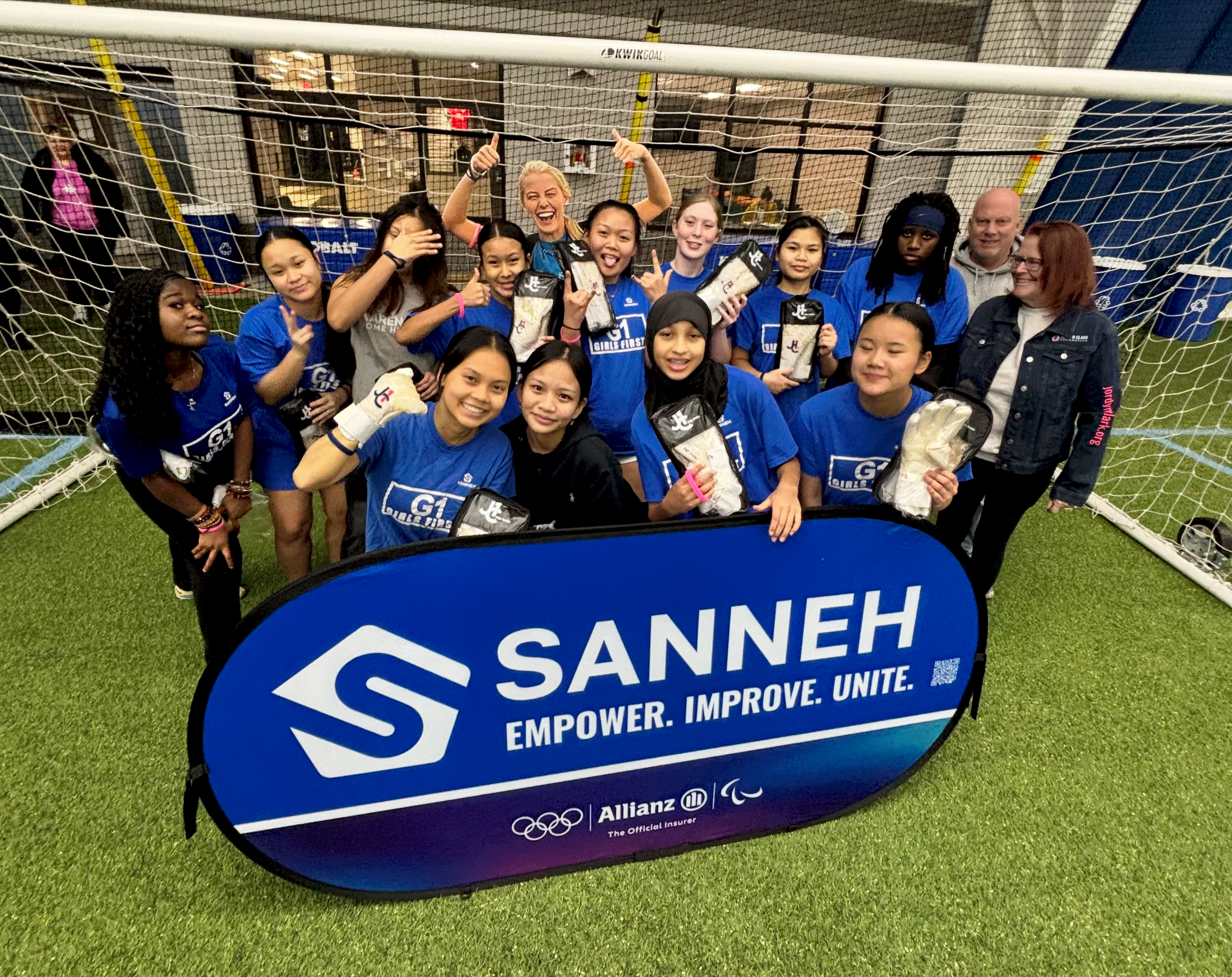 A group of coaches and girls standing behind a banner that says Sanneh, Empower, Improve, Unite.