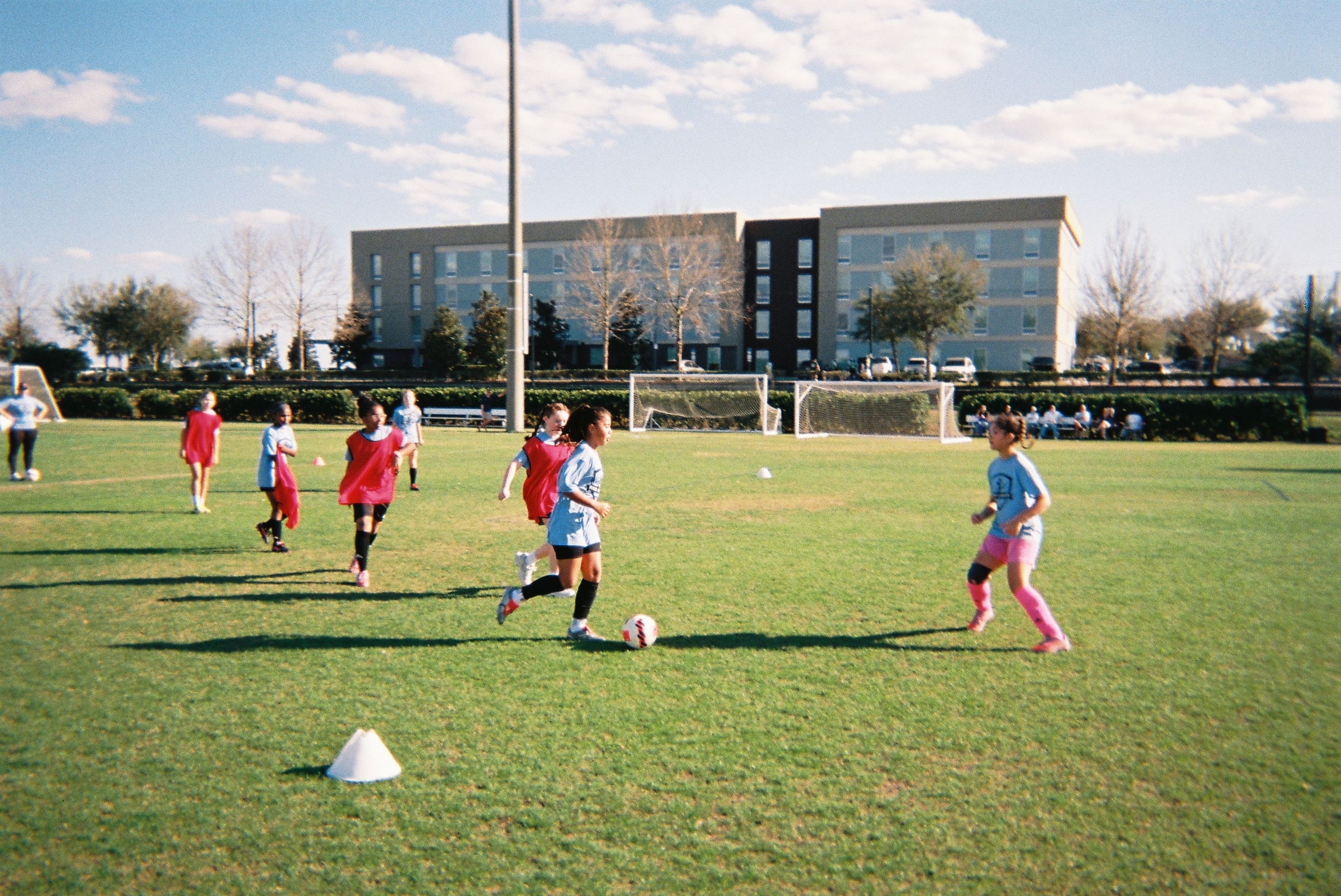 Girls play a small-sided soccer game on a grassy field, dribbling and defending around cones with goals in the background.