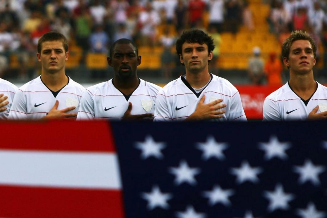 four usmnt players in white with a hand over their heart and a usa flag in the foreground