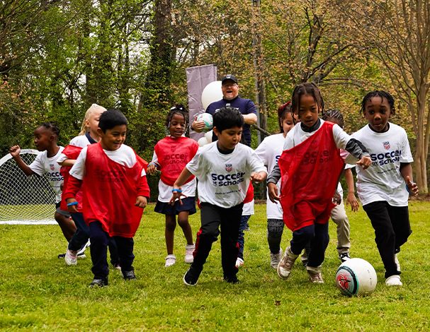 Young children playing soccer together outdoors during a Soccer at Schools session, chasing a ball across a grassy field.