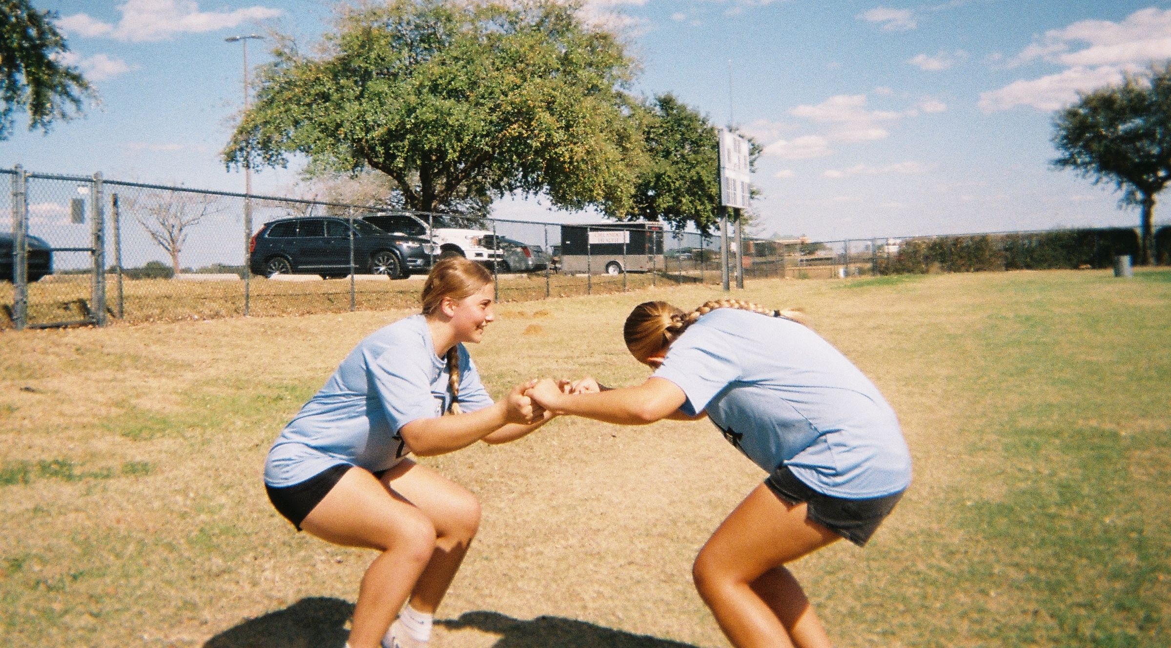 Two girls in team shirts crouch facing each other and hold hands during a partner exercise on a grassy field.