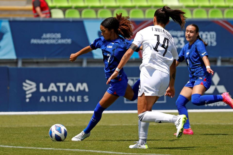 A US Player in blue takes a shot with a teammate trailing and opponent coming up to contest