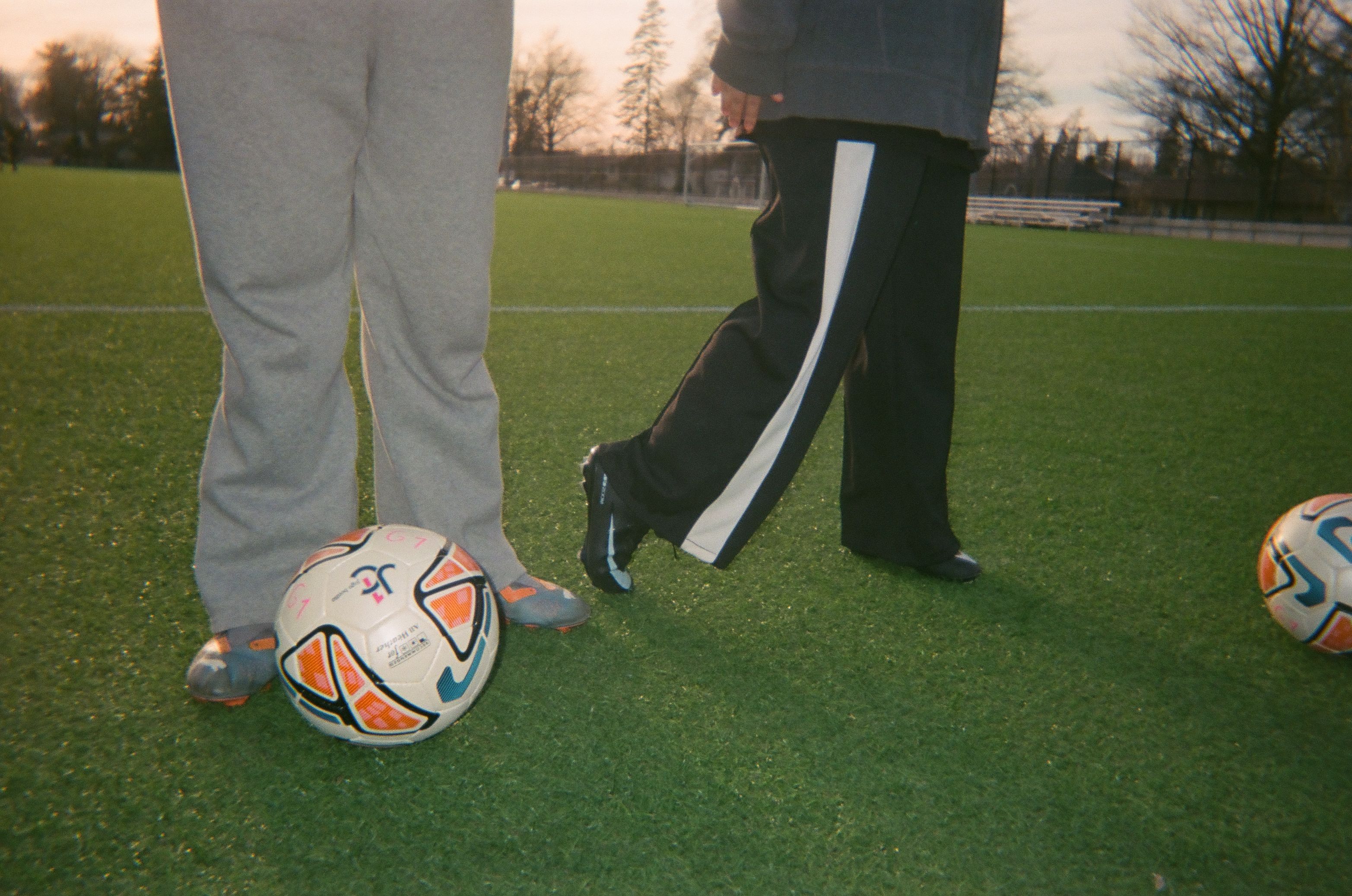 Two coaches standing with soccer balls at their feet.