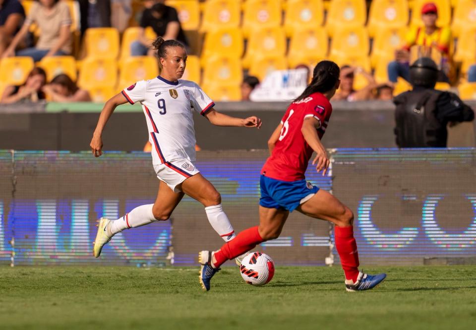 Mallory Swanson in a white jersey and shorts dribbles the ball towards a Costa Rican defender during a match