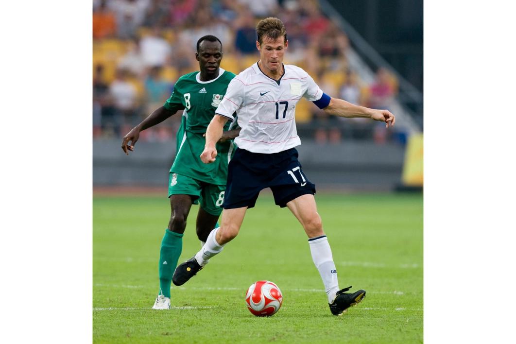 brian mcbride in white jersey and dark shorts dribbling a ball in front of an opponent