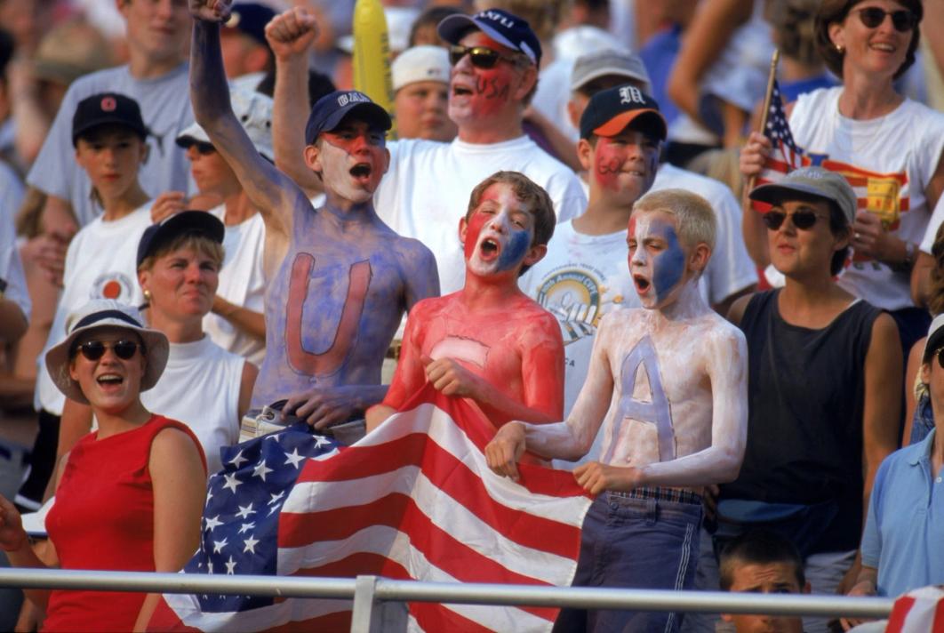 Three young male USA fans in blue, red and white body paint with letters USA holding a USA flag
