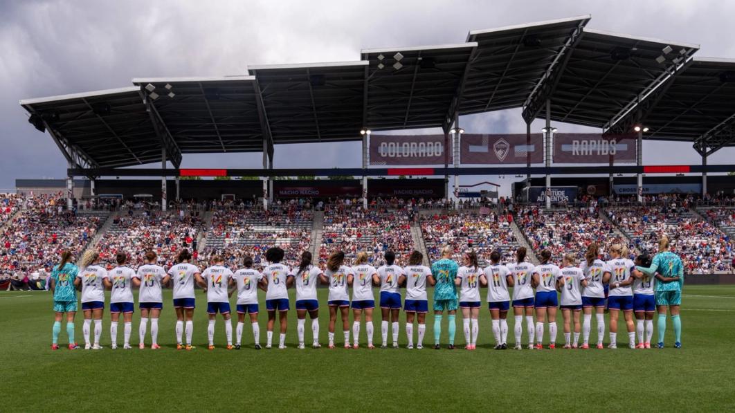 23 uswnt players with their backs facing the camera wearing white jerseys with progress flag colors