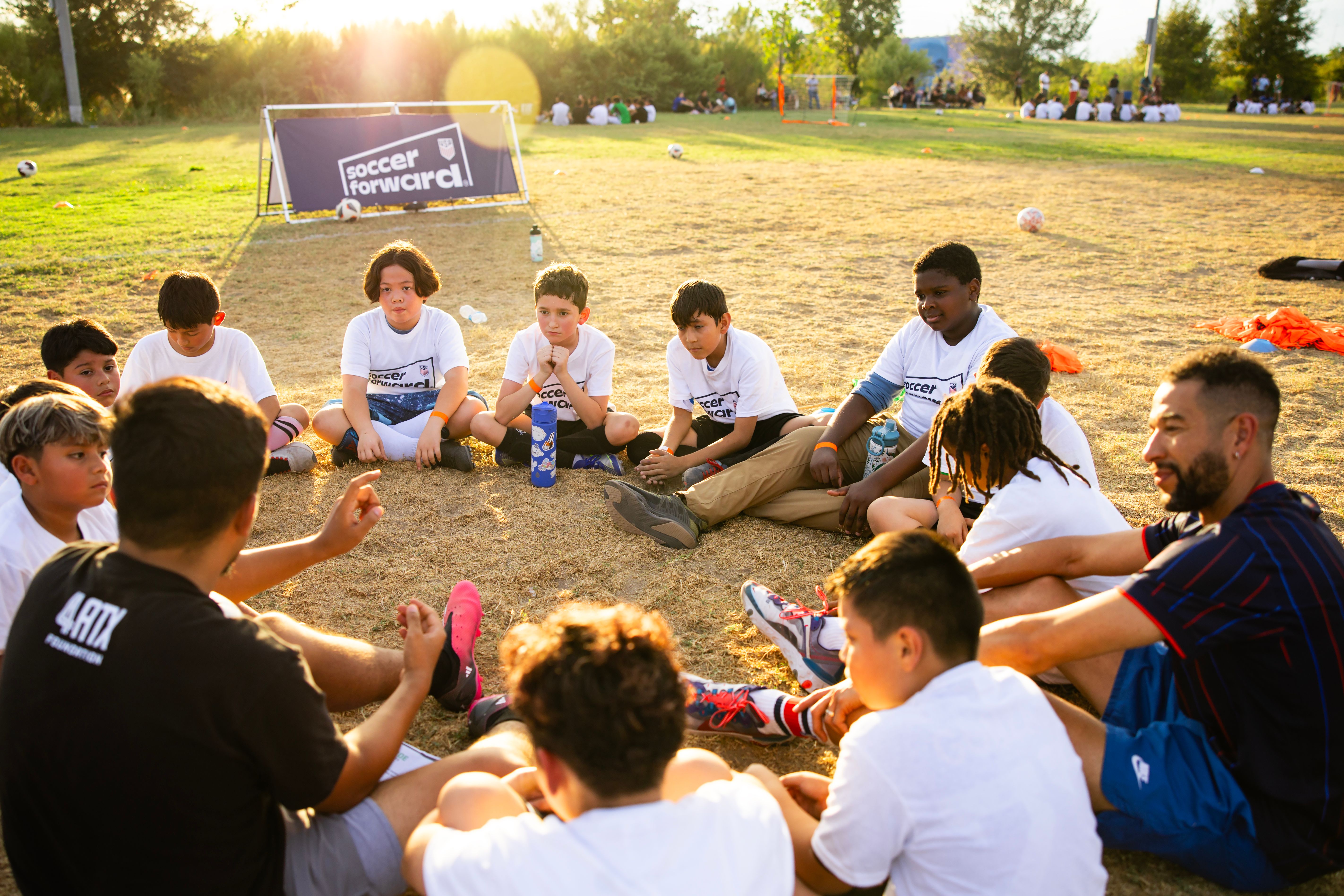 Coach and young player high-fiving on a soccer field during a training session, with other players nearby wearing bright pinnies.
