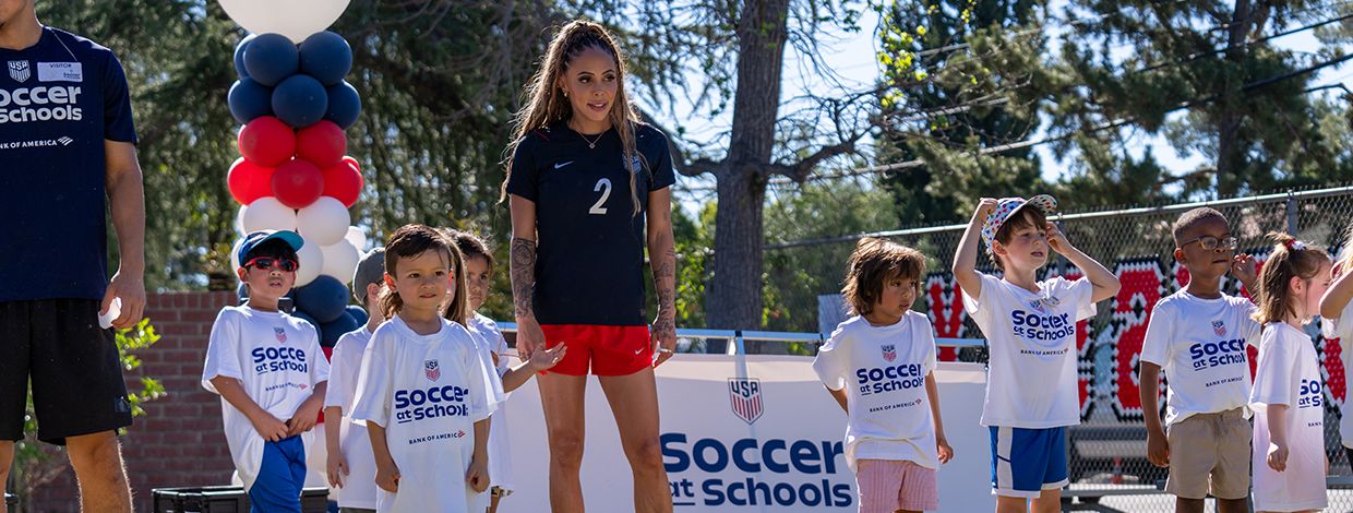 Children wearing Soccer at Schools shirts standing on a soccer field alongside a female soccer player during a community event.