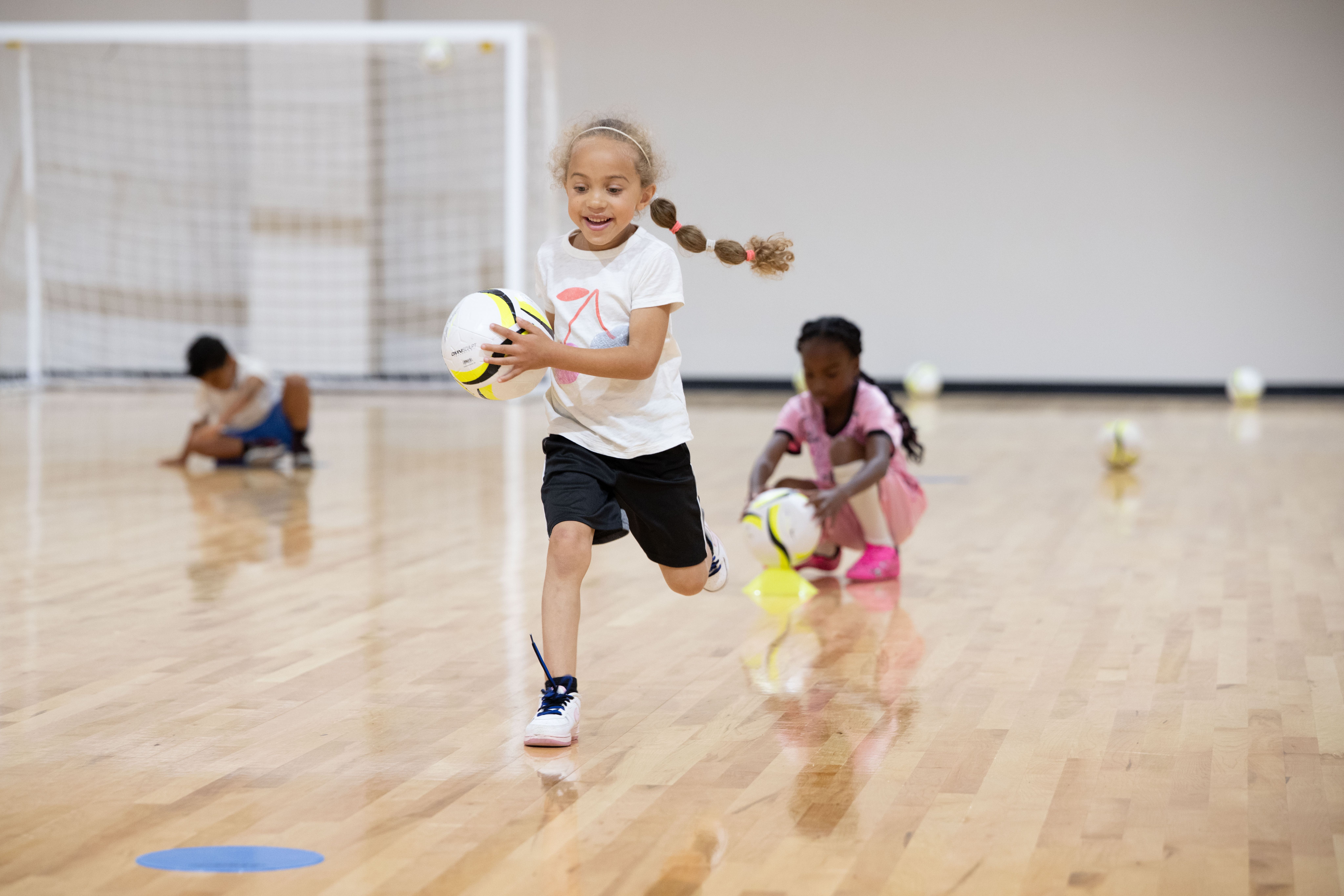 Children participating in an indoor soccer activity with soccer balls and cones.