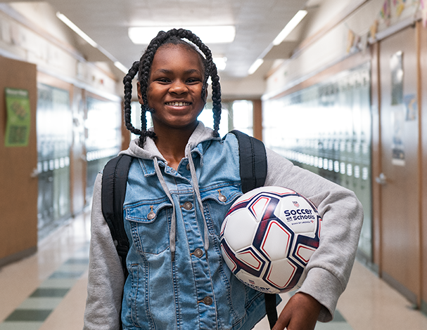 Smiling student holding a Soccer at Schools ball in a school hallway.