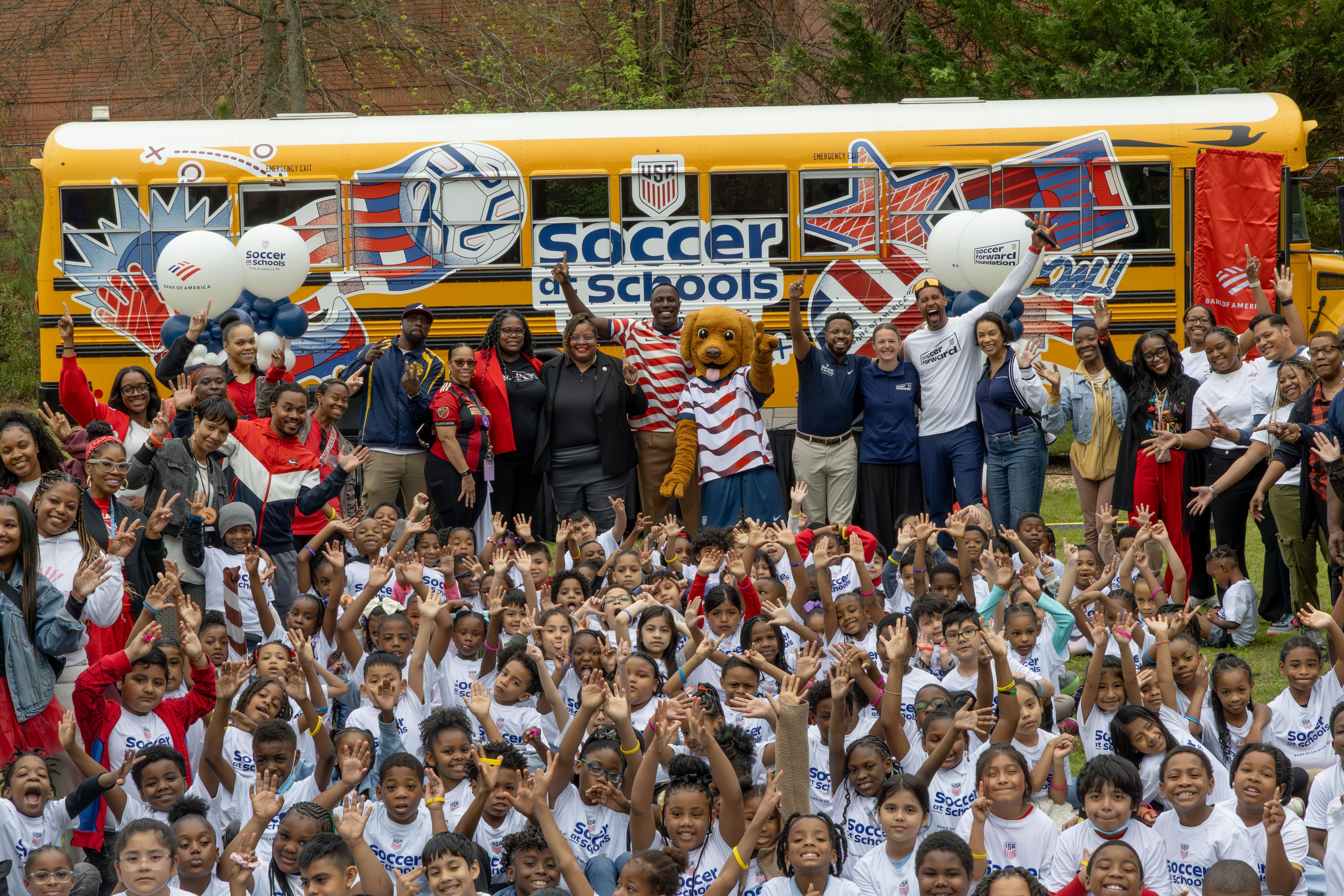 Students, educators, coaches, and event staff are gathered in front of a Soccer at Schools themed school bus with a large group of children in Soccer at Schools T-shirts. They are all smiling and celebrating the event day.