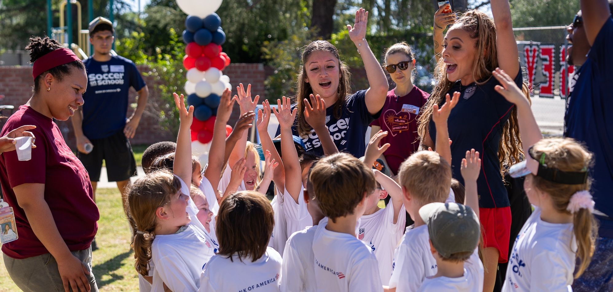 Children and coaches celebrate together with raised hands during a Soccer at Schools event outdoors.