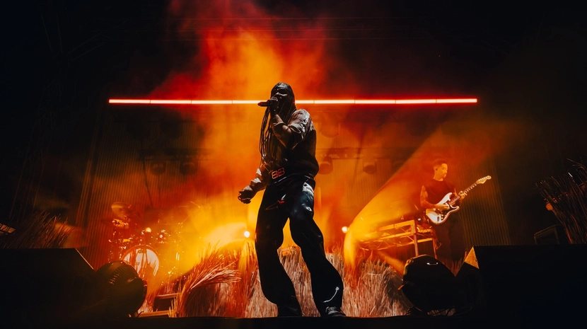 An artist performs on a dramatic stage filled with orange smoke and bright light, holding a microphone. To the right, a guitarist plays under a sharp beam of light against a backdrop of tall grass.