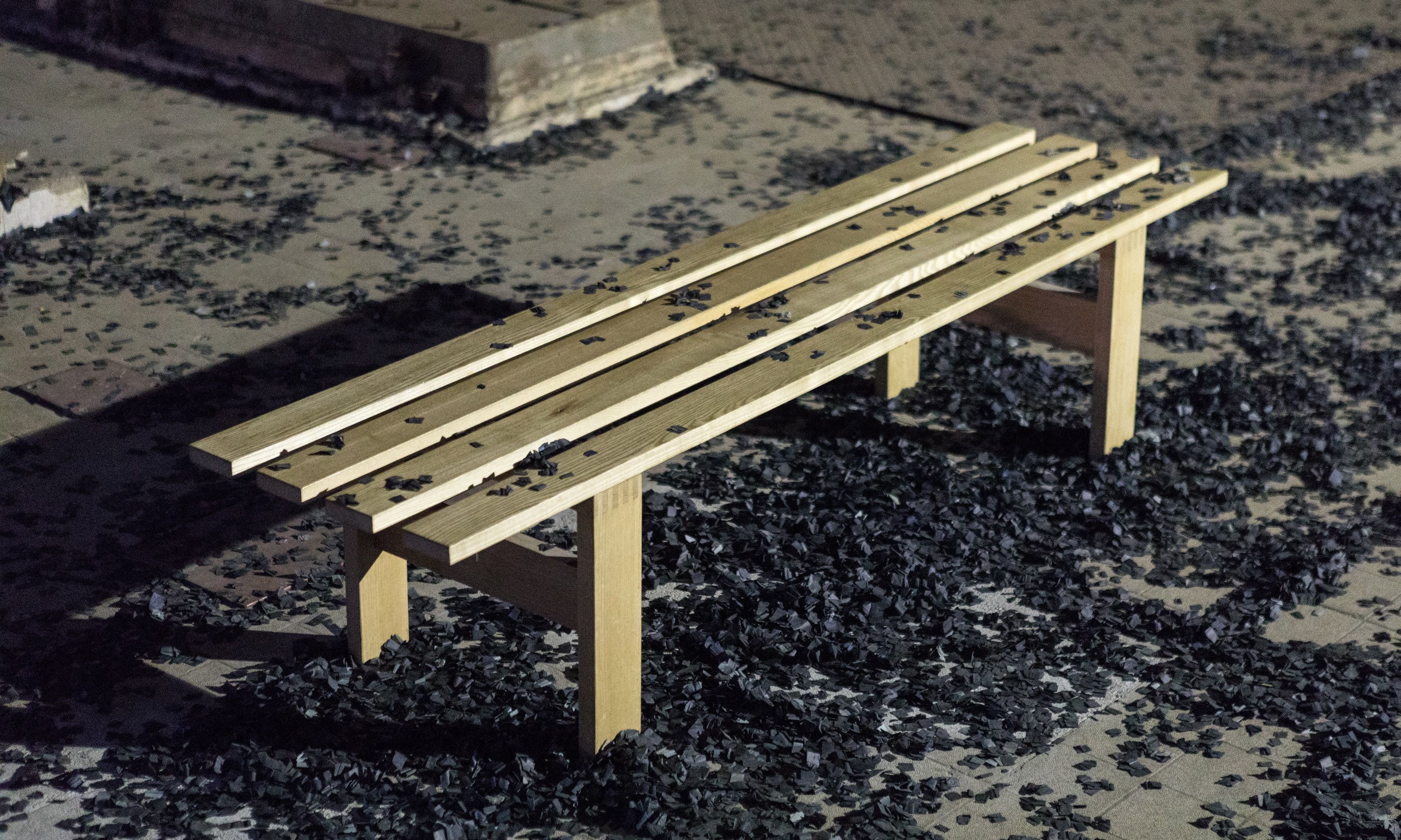 A simple light-wood bench sits outdoors, surrounded by a thick scattering of black, confetti-like debris on the ground. Some of the dark material also rests on the slats of the bench, creating a stark contrast against the wood.