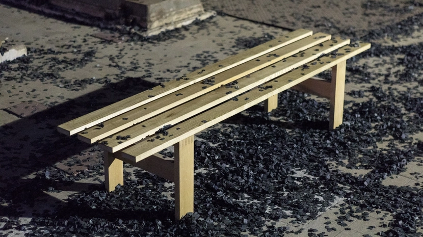 A simple light-wood bench sits outdoors, surrounded by a thick scattering of black, confetti-like debris on the ground. Some of the dark material also rests on the slats of the bench, creating a stark contrast against the wood.