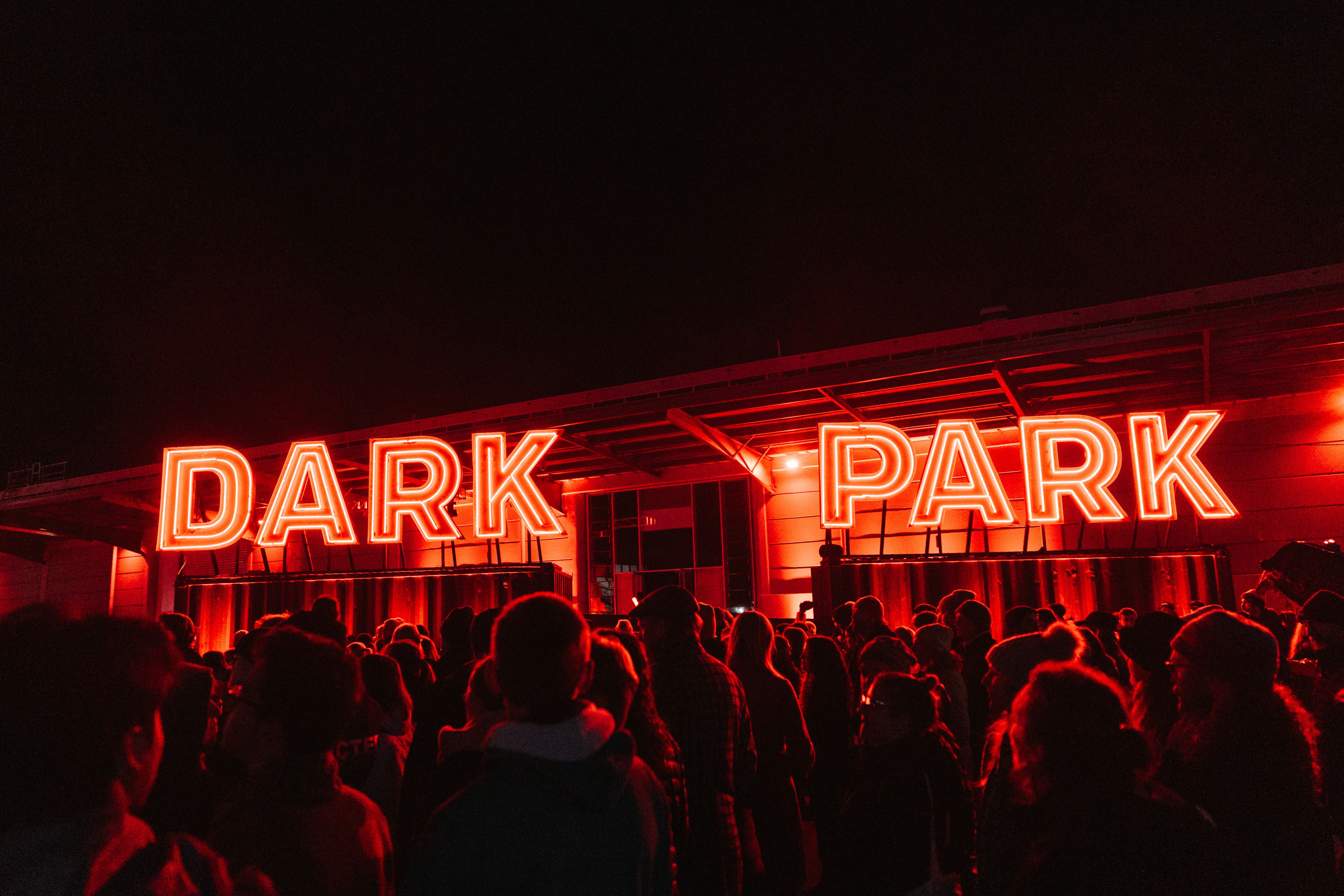 A large crowd of people gathered at night in front of a building featuring a glowing red neon sign that reads "DARK PARK." The entire scene is bathed in intense red light, contrasting against the pitch-black sky.