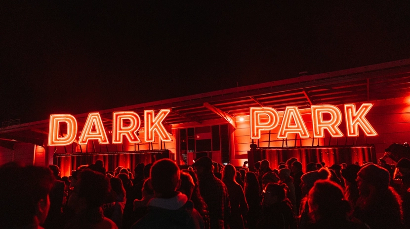 A large crowd of people gathered at night in front of a building featuring a glowing red neon sign that reads "DARK PARK." The entire scene is bathed in intense red light, contrasting against the pitch-black sky.