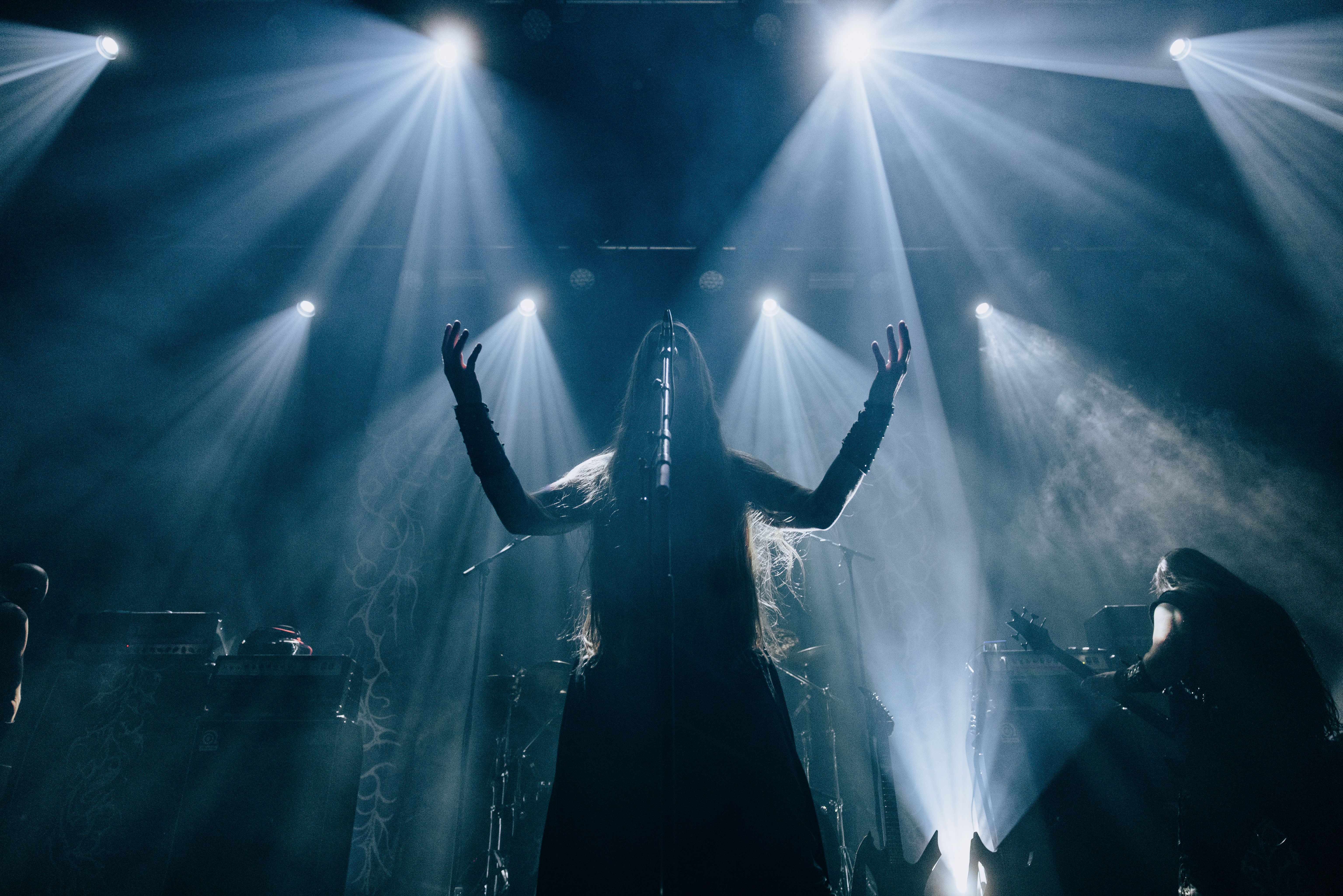 A dramatic silhouette of a long-haired performer with arms raised on a dark stage, illuminated by several sharp, radial beams of white light. A guitar player and stage equipment are visible in the misty, atmospheric background.