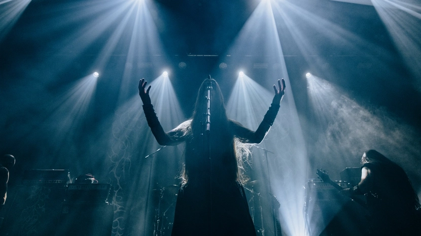 A dramatic silhouette of a long-haired performer with arms raised on a dark stage, illuminated by several sharp, radial beams of white light. A guitar player and stage equipment are visible in the misty, atmospheric background.