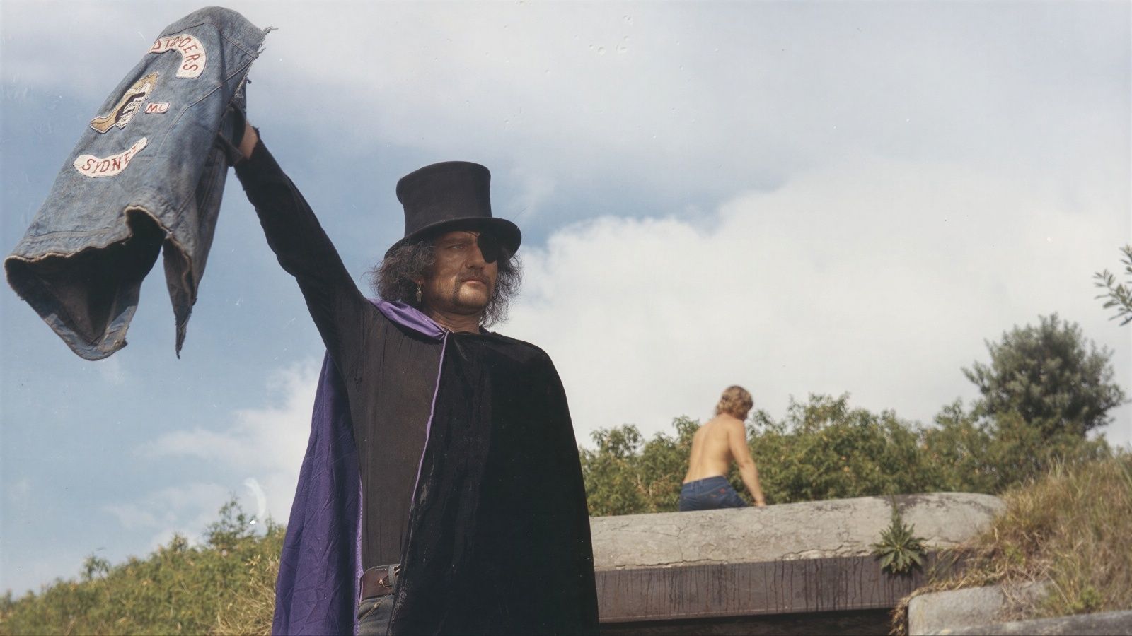 A man wearing a top hat, an eyepatch, and a purple-lined black cape holds a denim biker vest high in the air. He stands outdoors against a cloudy sky, with a shirtless man visible in the background atop a concrete structure.